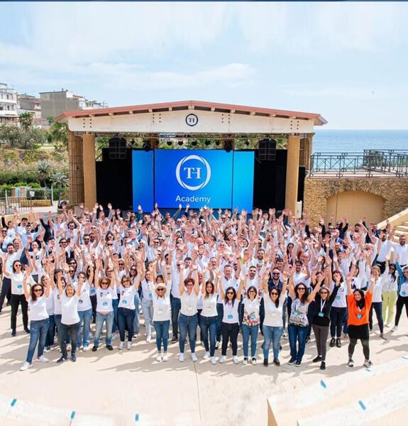 Group of people in white t-shirts gathered outdoors in front of a stage with a blue screen and TH Academy logo.