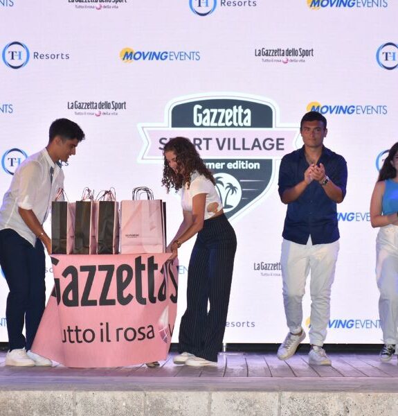 Four young people on stage during the Gazzetta Sport Village event with a table and pink gift bags.
