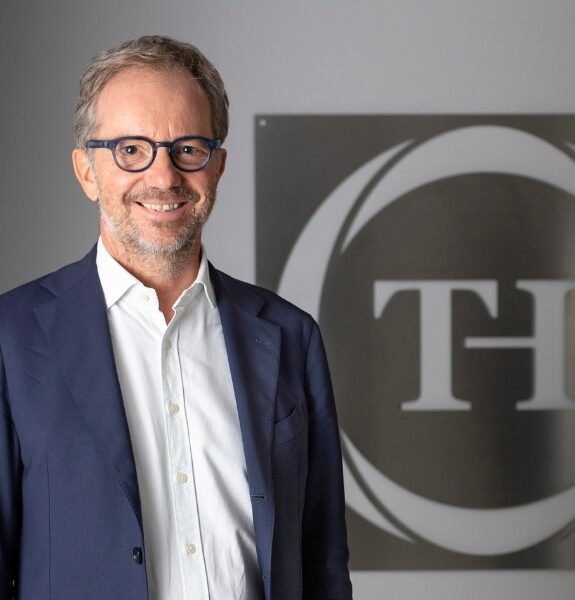 Man in a blue jacket and glasses, smiling, in front of a wall with a gray and white circular logo.