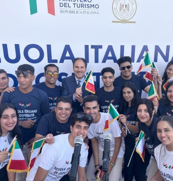 Group of young people wearing "Italian School of Hospitality" t-shirts holding Italian flags in front of a background featuring the logo of the Ministry of Tourism.