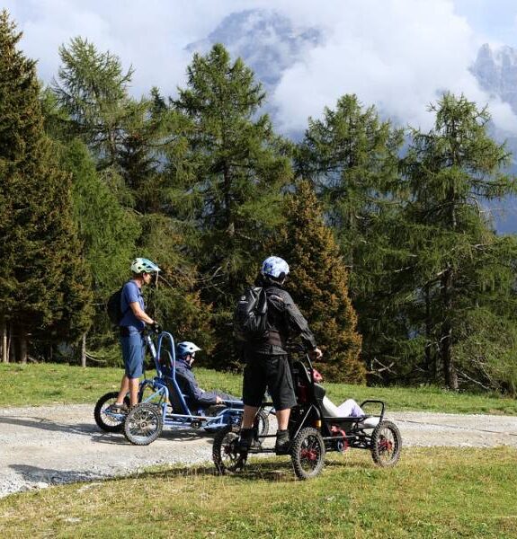 Two people in four-wheeled wheelchairs and two companions wearing helmets on a mountain path surrounded by green trees.