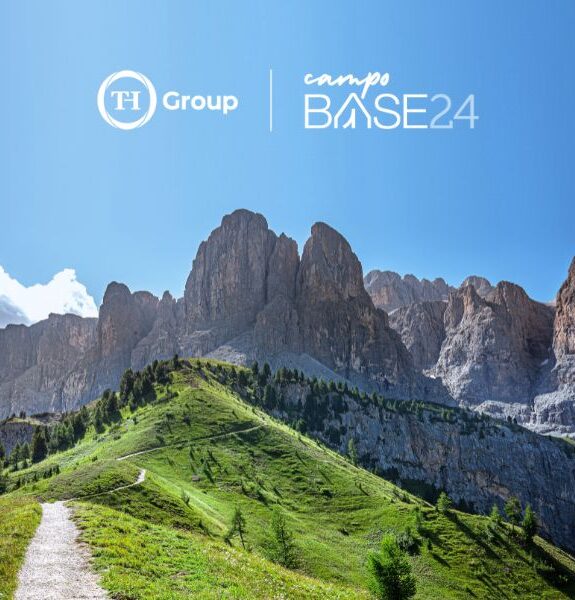 Mountain landscape with a path, green meadows, and high rocks under a clear sky, TH Group logo and Base Camp 24.