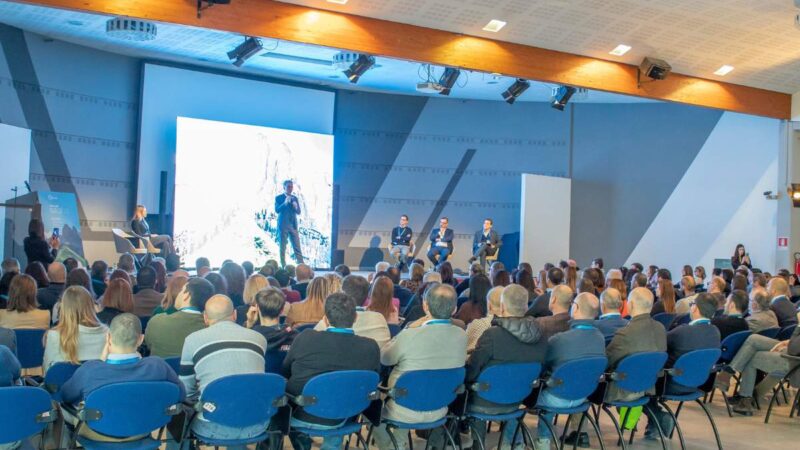 Conference with a large audience in the room, speakers on stage, and a big screen illuminated in the background.