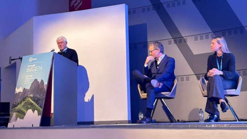 Three people on stage at a conference, with a man speaking at the podium and two seated listeners, professional context.