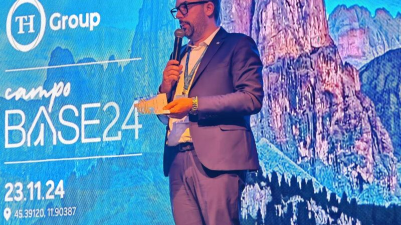 Man with a microphone and notes speaks in front of a screen with the TH Group logo and a mountain landscape.