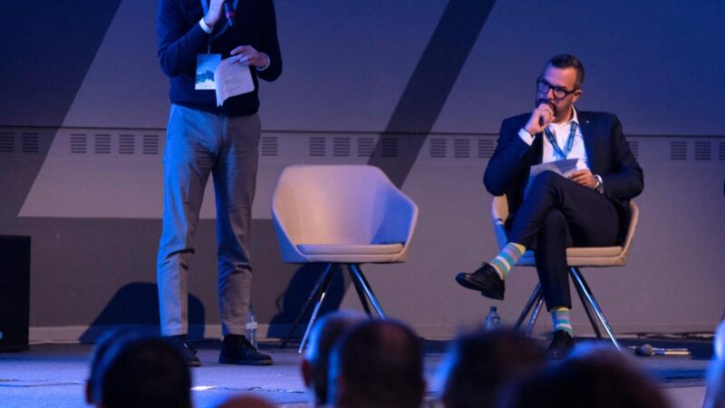 A man standing with a microphone speaks at a conference, another man sitting listens thoughtfully, the audience visible in the foreground.