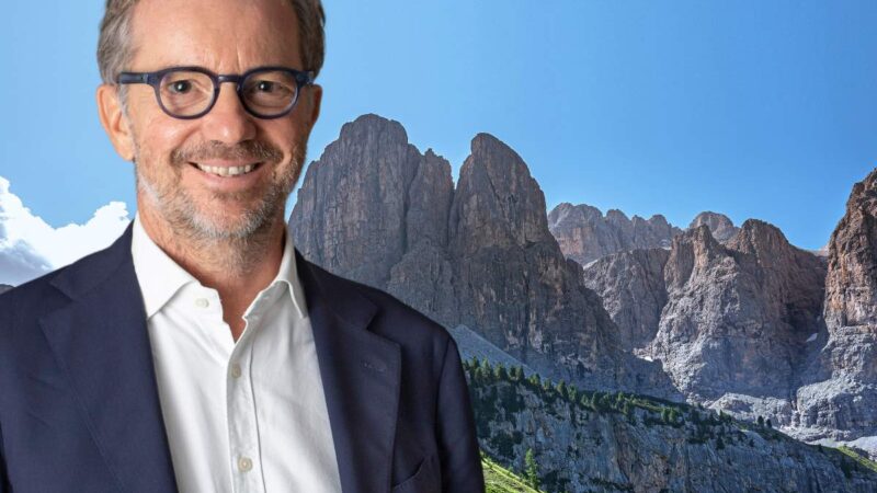 Portrait of a smiling man with glasses and a jacket, against a background of rocky mountains and a light blue sky.