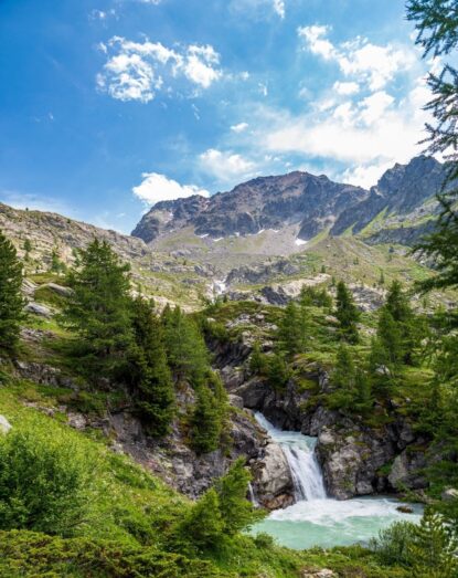 Cascata di montagna tra rocce e alberi verdi, cielo azzurro con nuvole sparse.