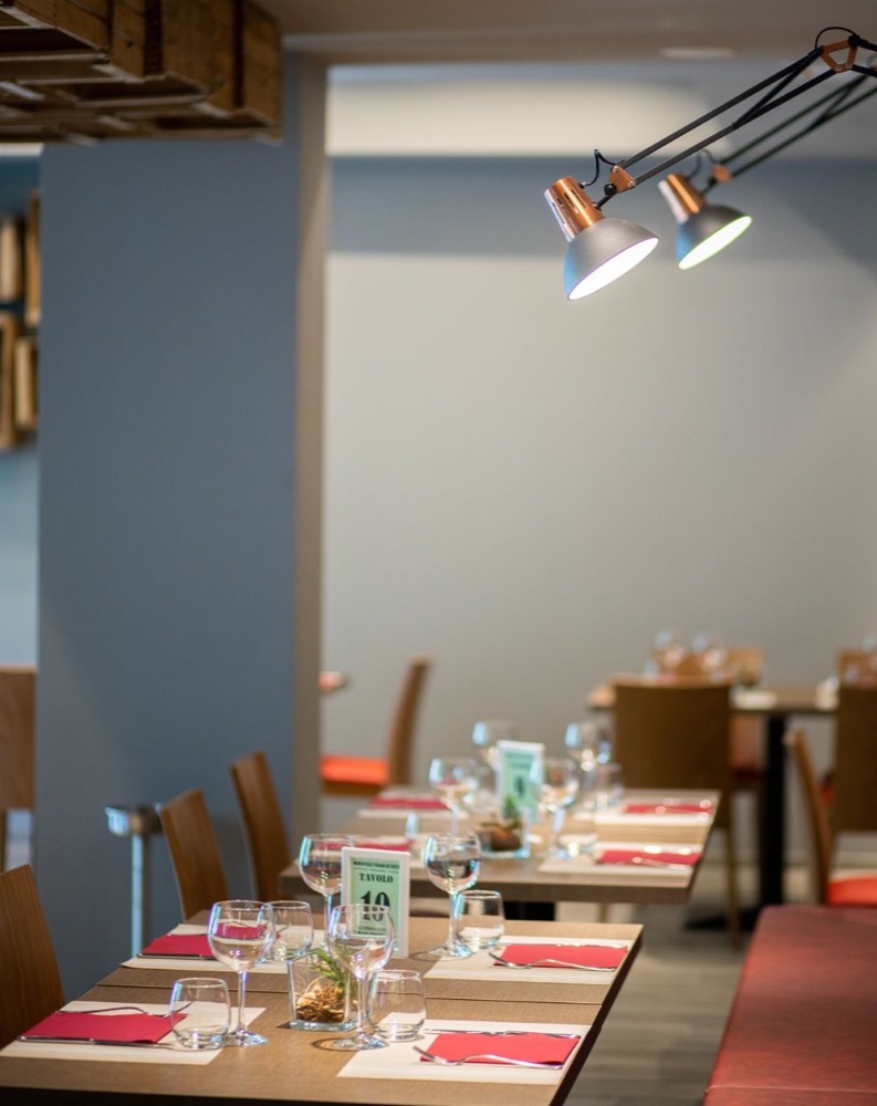 Interno di ristorante con tavoli preparati, bicchieri e posate, illuminazione diretta sopra i tavoli.