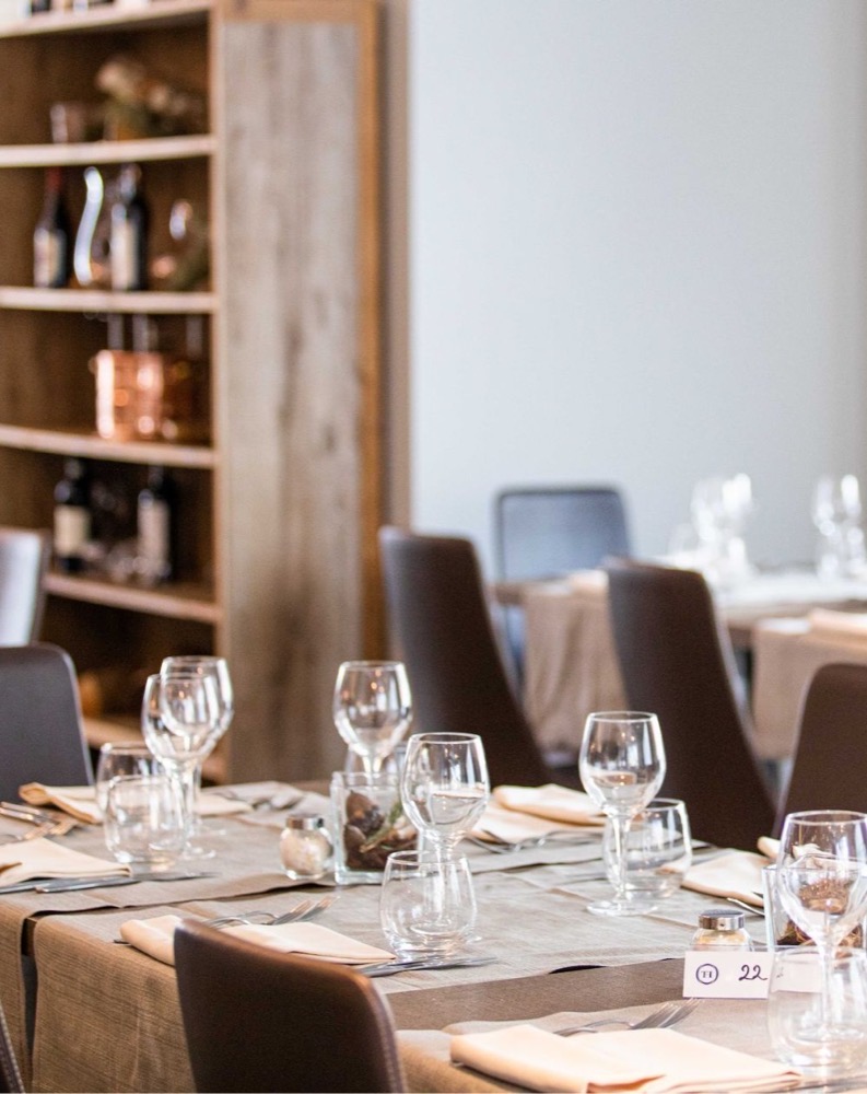 Tavola apparecchiata con bicchieri, posate e tovaglioli in un ristorante elegante, sfondo con scaffale di bottiglie e oggetti decorativi.