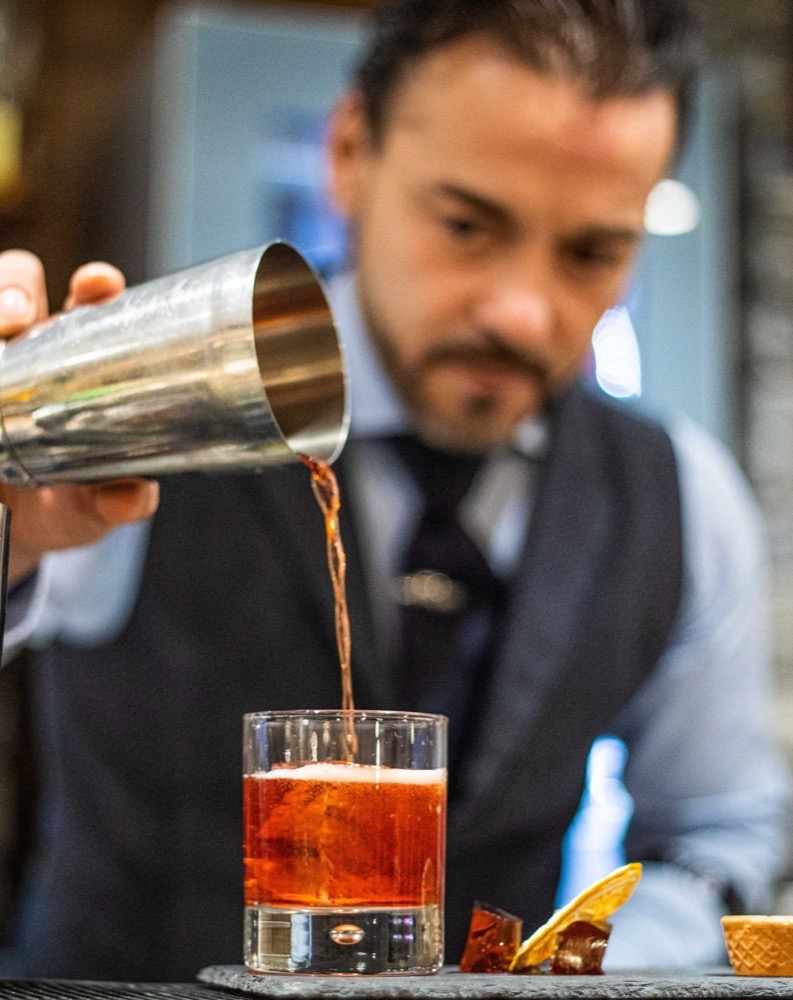 Barman versa un cocktail rosso in un bicchiere trasparente, con fette di limone e ghiaccio sul bancone.