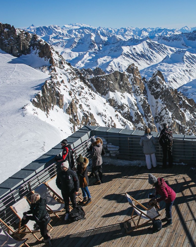 Persone che ammirano la vista montana innevata da una terrazza panoramica, con cime e valli circostanti.