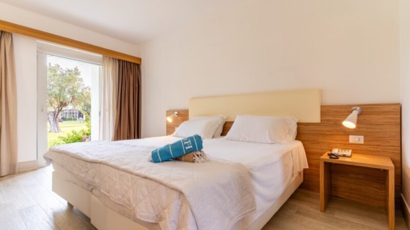 Camera da letto luminosa con letto matrimoniale, asciugamani blu arrotolati, comodino in legno e finestra con vista sul giardino.