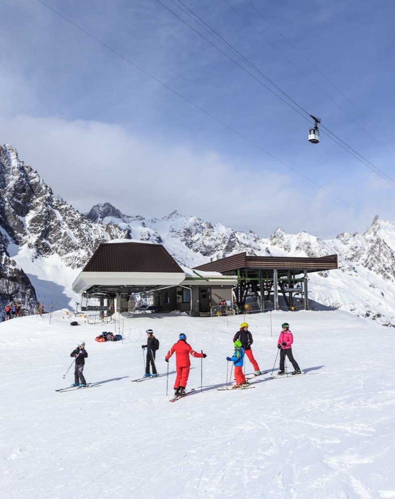 Gruppo di sciatori bambini e adulti fuori dalla funivia in montagna con neve e cime innevate sullo sfondo.