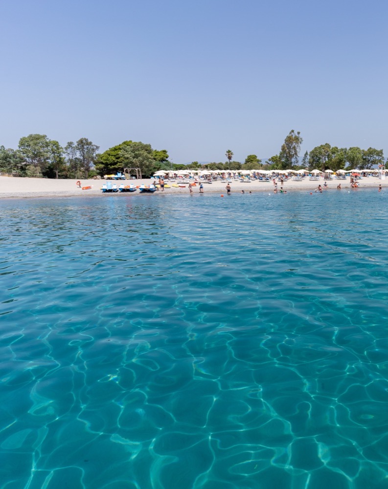 Spiaggia affollata con ombrelloni e persone, vista dal mare con acqua limpida e cielo sereno.