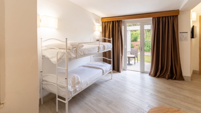 Camera con letto a castello bianco, pavimento chiaro, tende marroni e porta finestra che dà su un giardino.