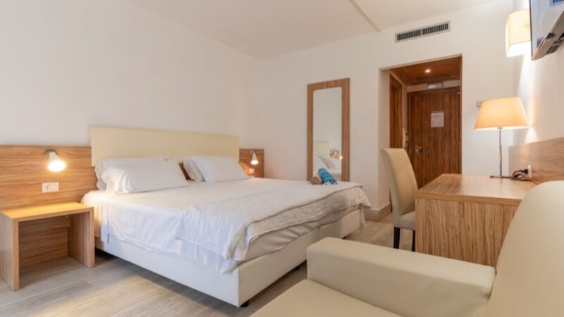 Camera d’albergo luminosa con letto matrimoniale, scrivania in legno, sedia beige e specchio a figura intera.