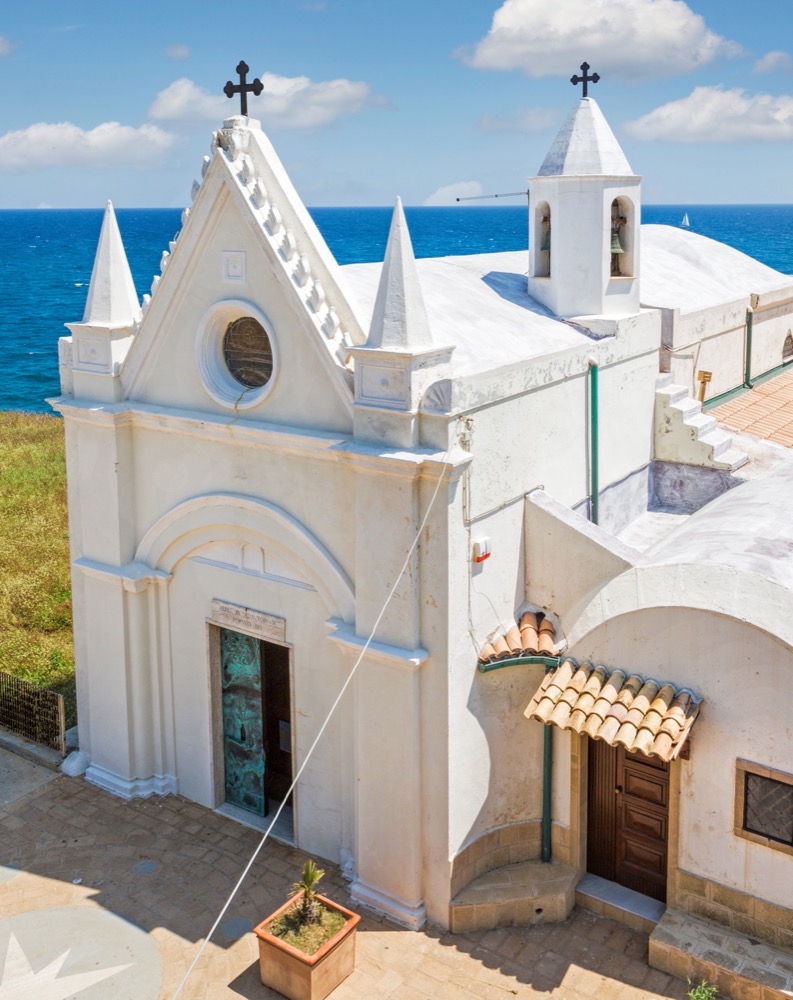 Chiesa bianca con campanile e croce sul tetto, sullo sfondo il mare azzurro e cielo sereno.