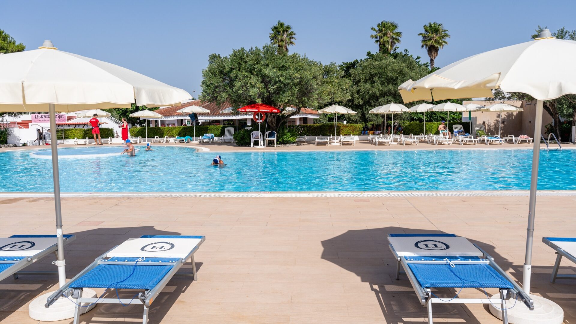 Basen na świeżym powietrzu z niebieskimi leżakami, białymi parasolami i ludźmi pływającymi w słoneczny dzień.