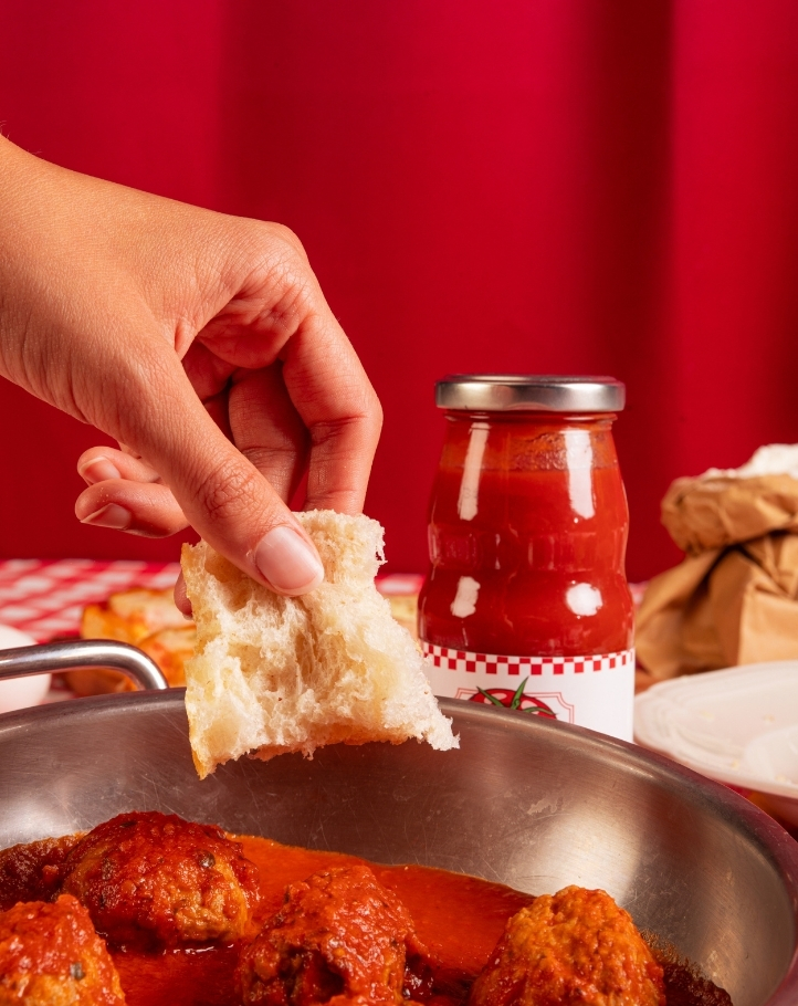 Una mano tiene un pezzo di pane mentre intinge in una pentola con polpette e salsa, con una bottiglia di salsa accanto.
