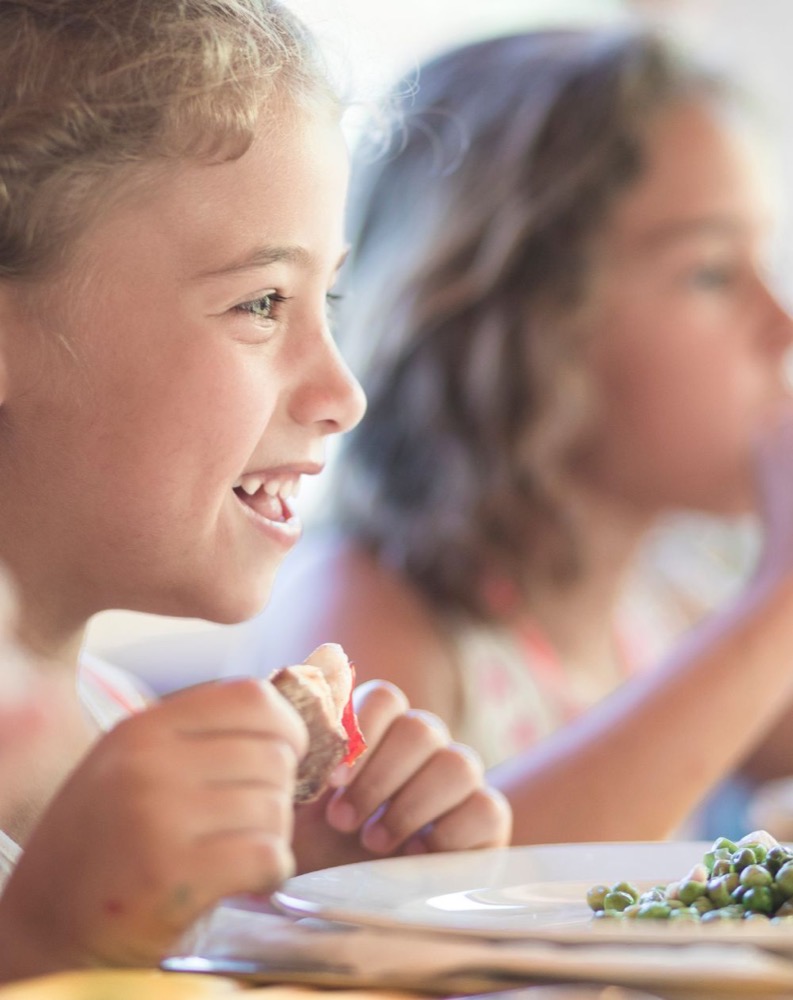 Ragazza sorridente mentre mangia, con un piatto di piselli in primo piano e altri bambini sullo sfondo.