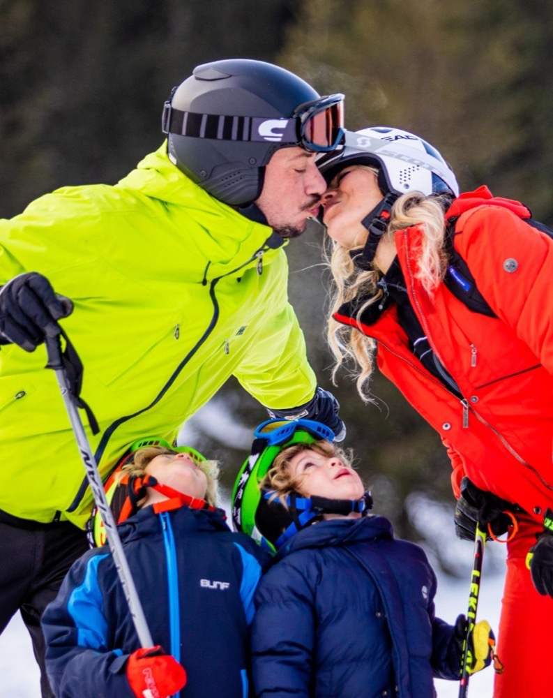 Una coppia si bacia in montagna, mentre due bambini li guardano ammirati, vestiti in tute e caschi da sci colorati.