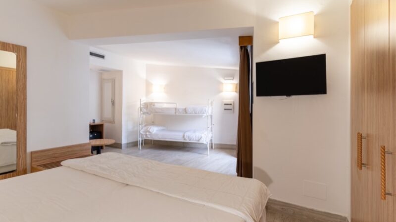 Camera luminosa con letto matrimoniale, letto a castello bianco, TV a parete e armadio in legno chiaro.