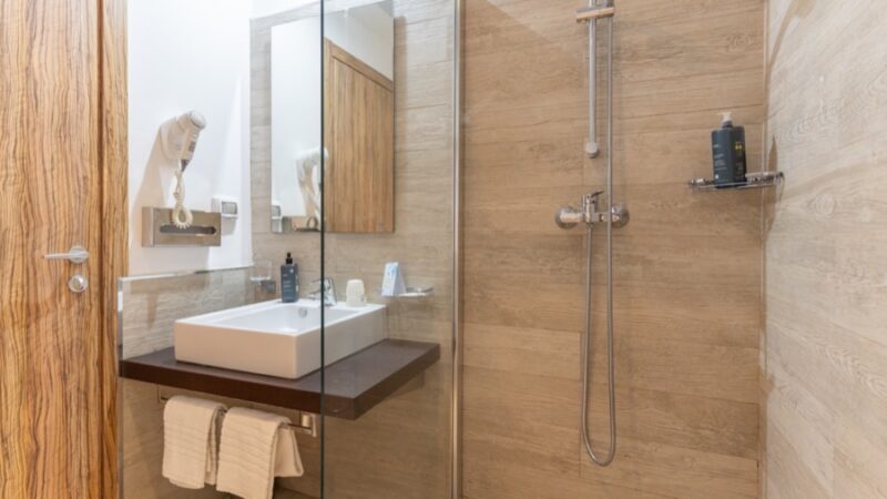 Bagno moderno con lavabo quadrato su piano scuro, doccia con parete in vetro e pareti effetto legno chiaro.