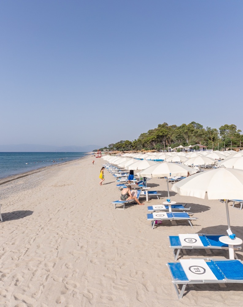Spiaggia sabbiosa con file di ombrelloni bianchi e lettini blu vicino al mare calmo sotto cielo sereno.
