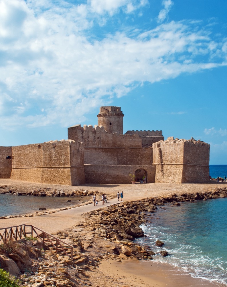 Castello medievale su isola rocciosa collegata alla terraferma da passerella con persone e mare azzurro.