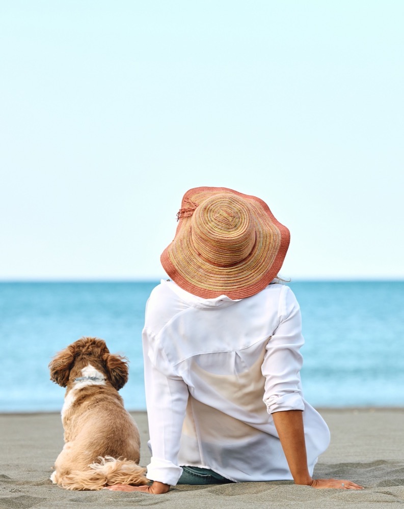 Donna seduta sulla spiaggia con il suo cane, indossa un cappello largo e una camicia bianca.