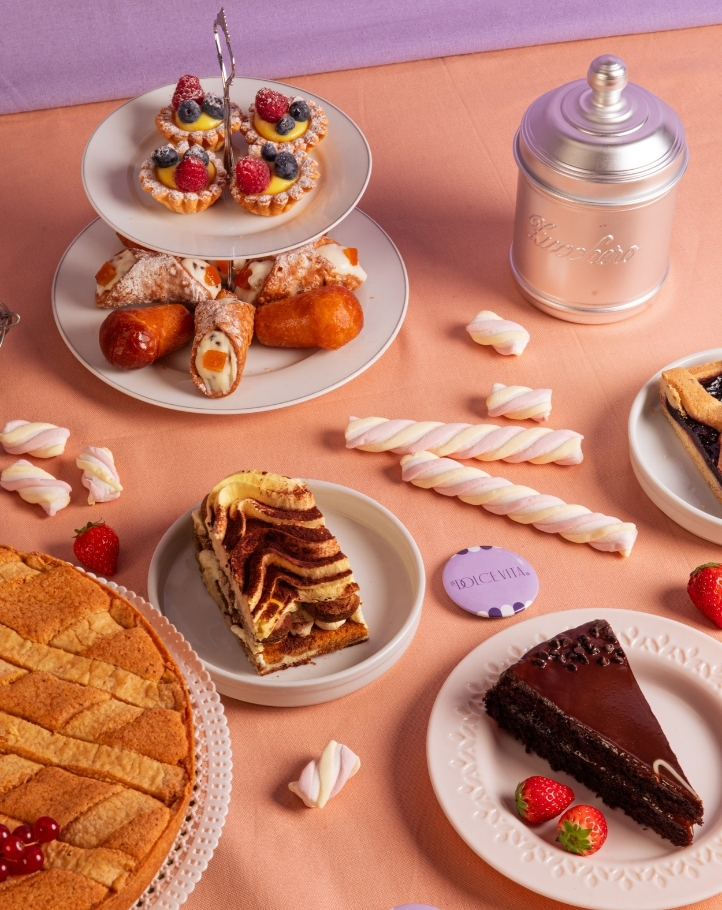 Tavolo imbandito con dolci: torta a fetta, crostate e cannoli, decorato con fragole e marshmallow su sfondo colorato.