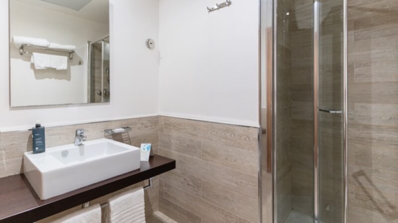 Bagno moderno con lavabo rettangolare su ripiano scuro, specchio, doccia con ante in vetro e piastrelle color legno.