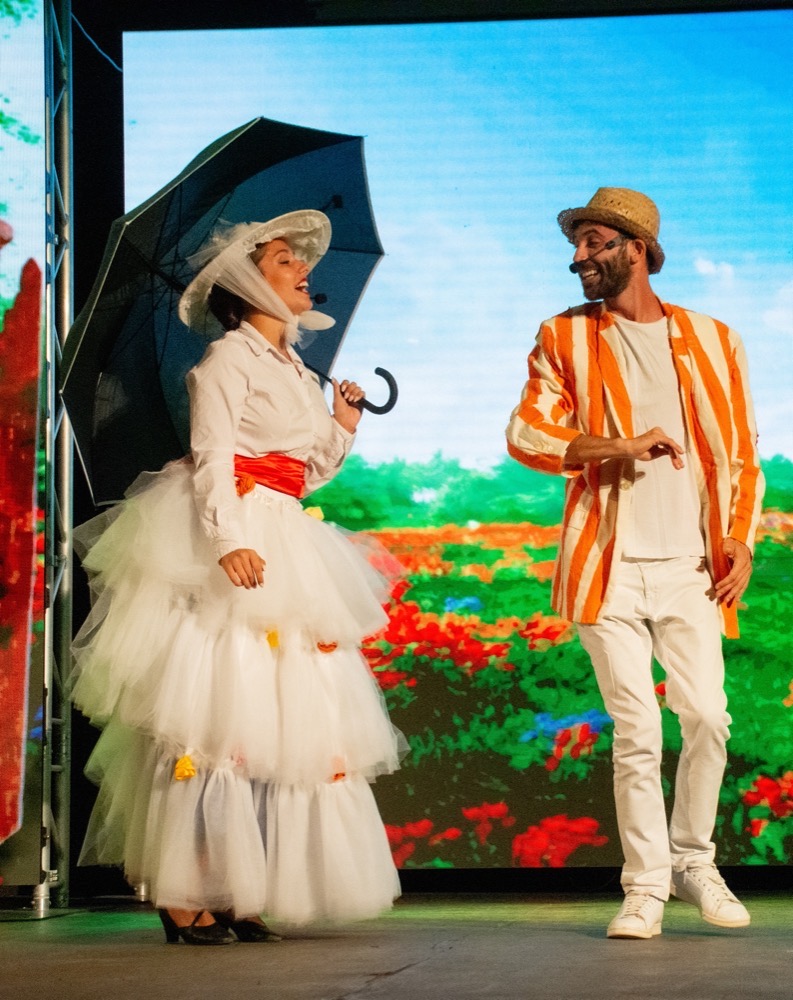 Due attori in costume colorato su un palco con sfondo floreale, lei con ombrello e lui con cappello di paglia.