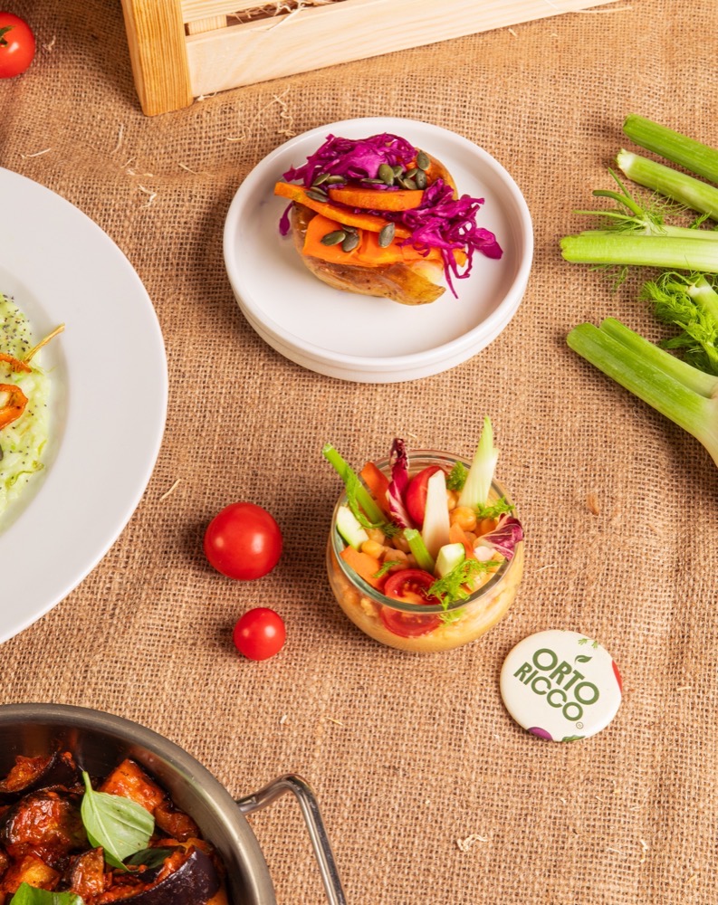 Insalata mista con pomodori, cetrioli e verdure, accompagnata da un piatto di verdure rosolate, su una superficie di stoffa.