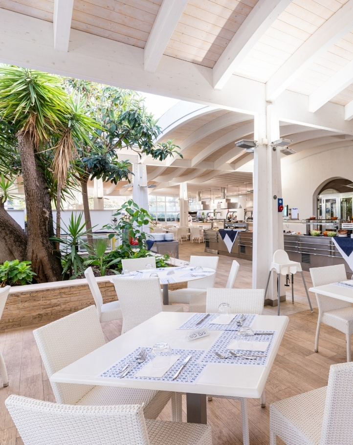 Interno di un ristorante elegante con tavoli bianchi, piante verdi e cucina visibile sullo sfondo. Atmosfera luminosa e accogliente.