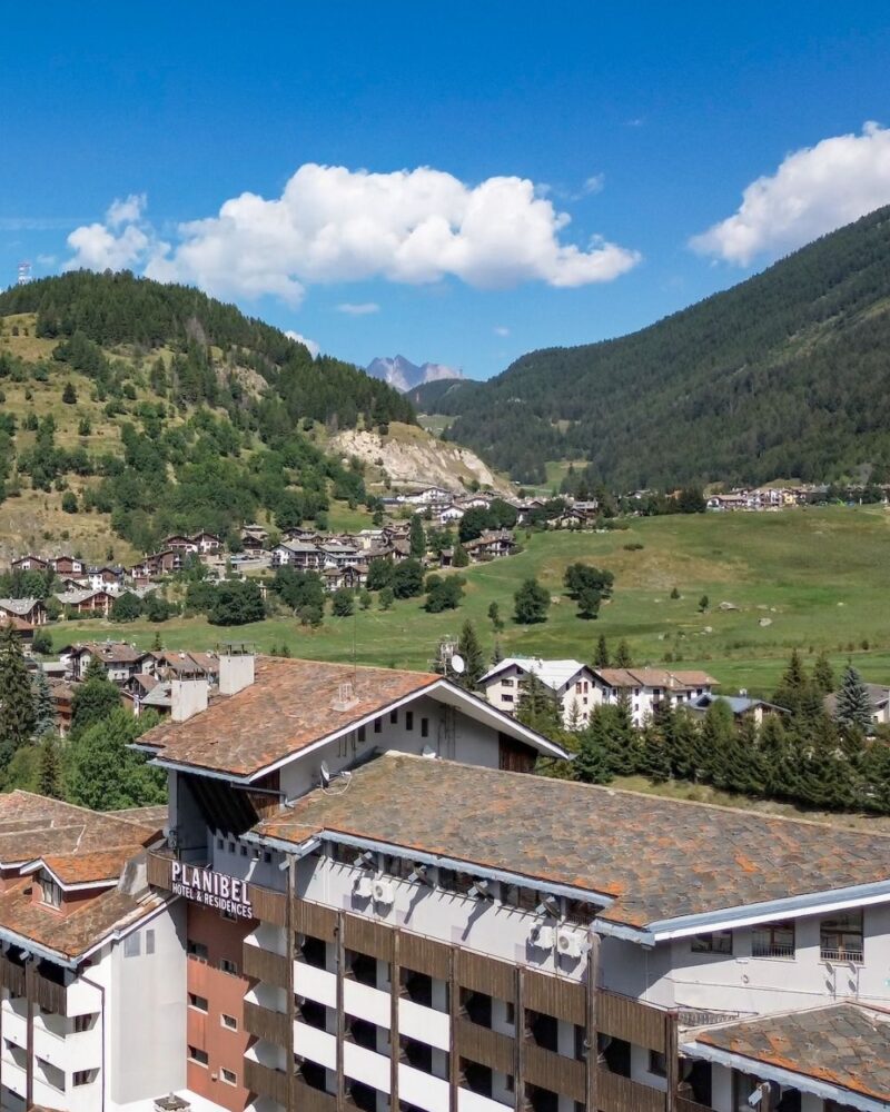 Paesaggio montano con alberghi, prati verdi e boschi sotto un cielo azzurro con alcune nuvole bianche.