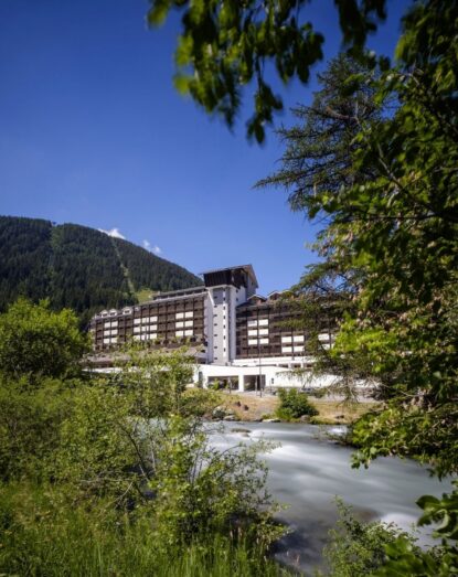 Albergo bianco di montagna vicino a un ruscello, circondato da alberi verdi e un cielo azzurro limpido.