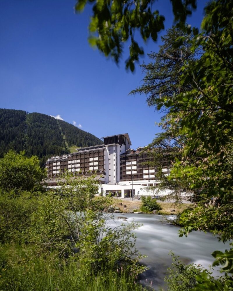 Albergo bianco di montagna vicino a un ruscello, circondato da alberi verdi e un cielo azzurro limpido.