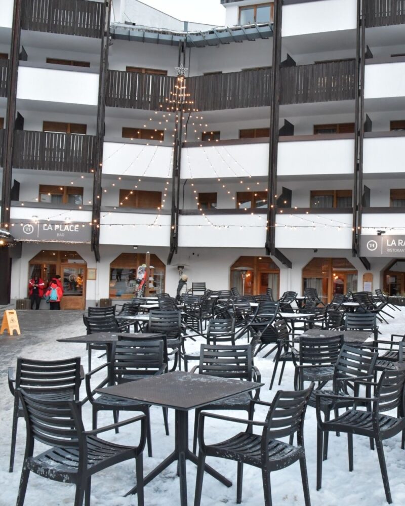 Cortile invernale con sedie e tavoli neri su neve davanti a un edificio bianco con balconi e luci a forma di albero.