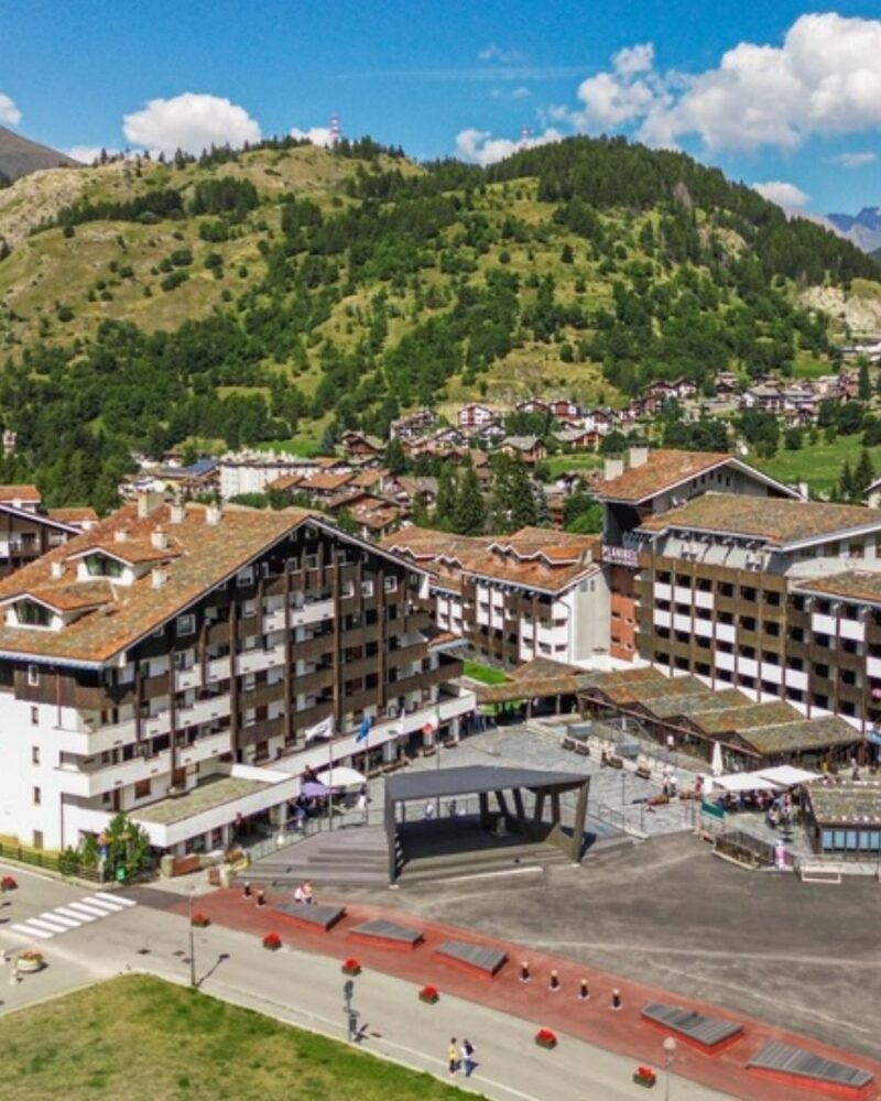 Vista panoramica di un paese alpino con edifici tradizionali, piazza centrale e colline verdi sullo sfondo.