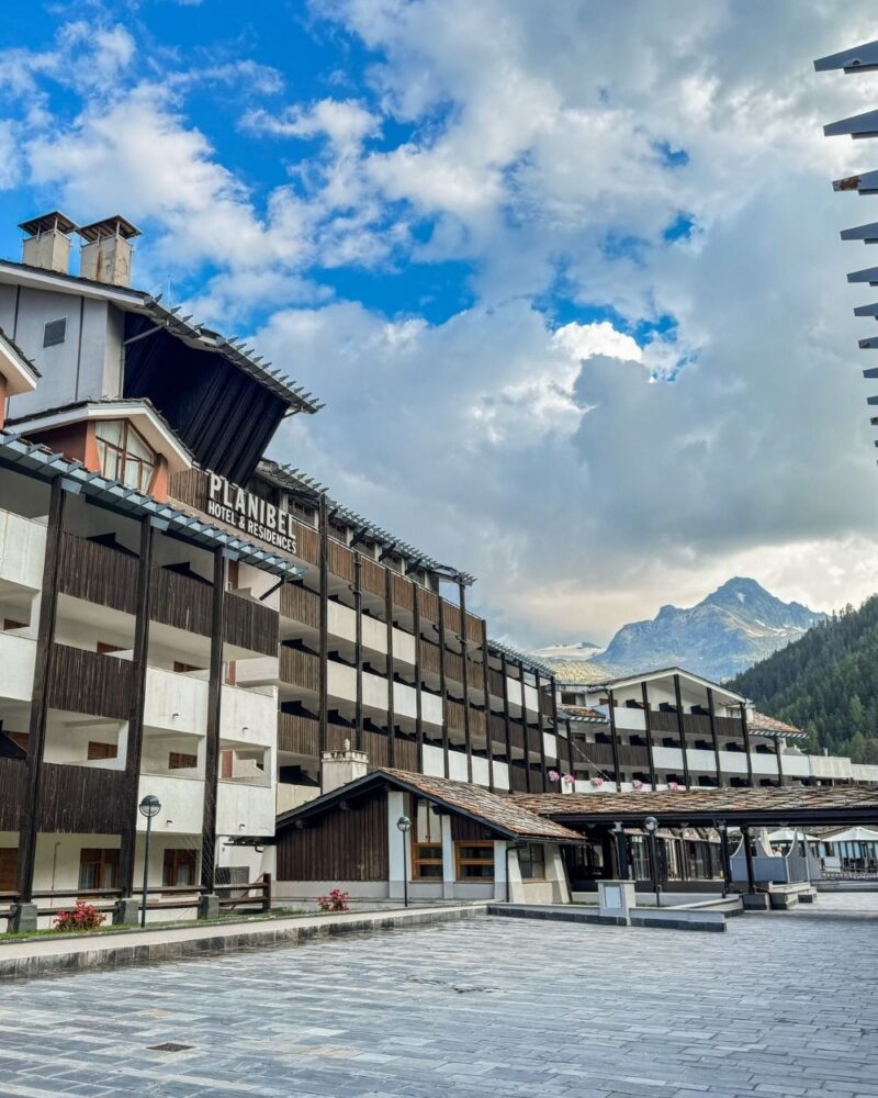 Hotel Planibel con balconi in legno sotto un cielo nuvoloso, con montagne e foreste sullo sfondo.