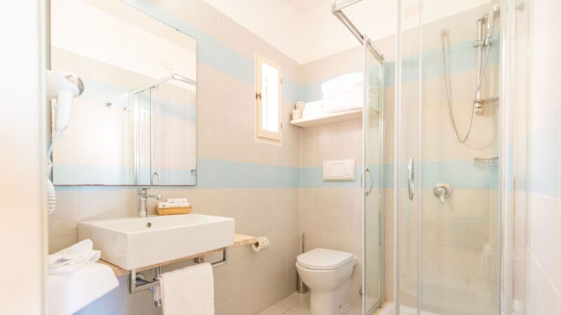 Bagno luminoso con lavabo, specchio, WC, doccia in vetro e scaffale con asciugamani bianchi.