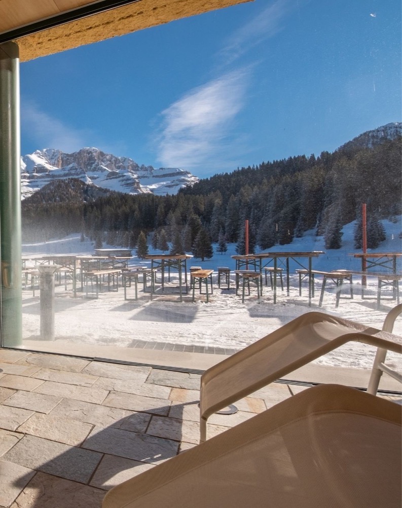 Vista panoramica delle montagne innevate e degli alberi dal bordo di una terrazza con sedie all'aperto.