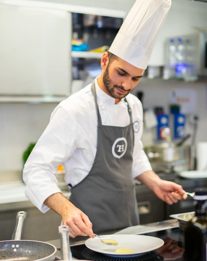 Chef in cucina con cappello, che prepara un piatto con componenti gialle.