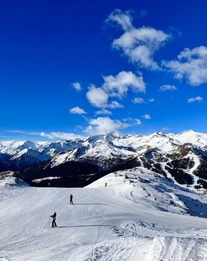 Sciatori su una pista innevata con montagne innevate e cielo blu con nuvole.