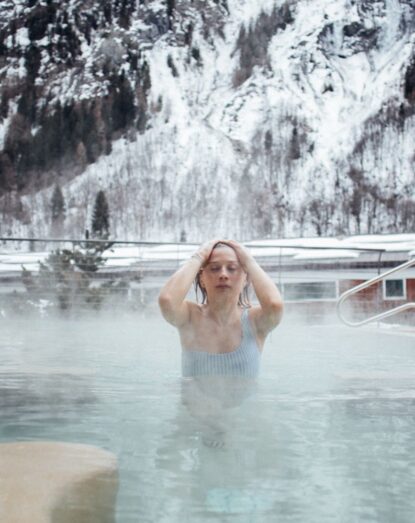 Woman in an outdoor pool with snow-capped mountains in the background, warm water and visible steam.