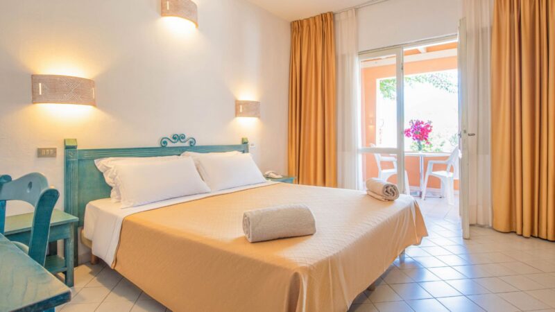 Camera da letto luminosa con letto matrimoniale, asciugamani arrotolati, mobili verdi e porta finestra che dà su balcone con fiori rosa.