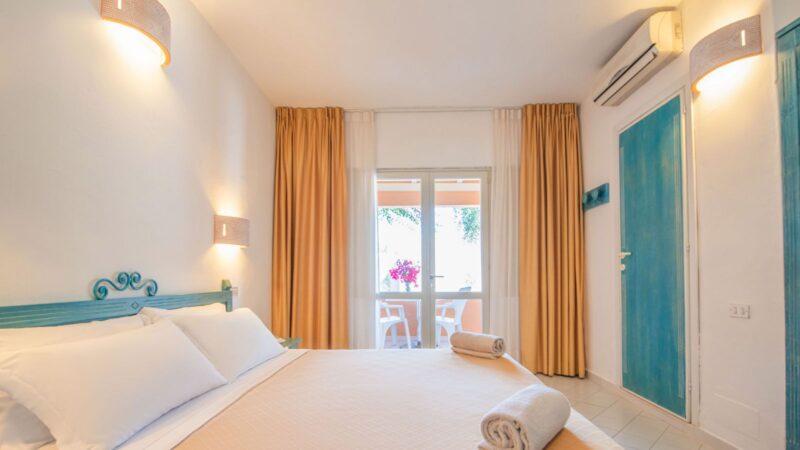 Camera da letto luminosa con letto matrimoniale, tende arancioni, porta finestra e balcone con sedie bianche e fiori rosa.