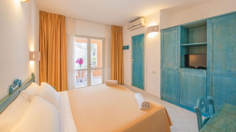 Camera da letto con letto matrimoniale, armadio azzurro, tende arancioni e porta finestra che si apre su un balcone.